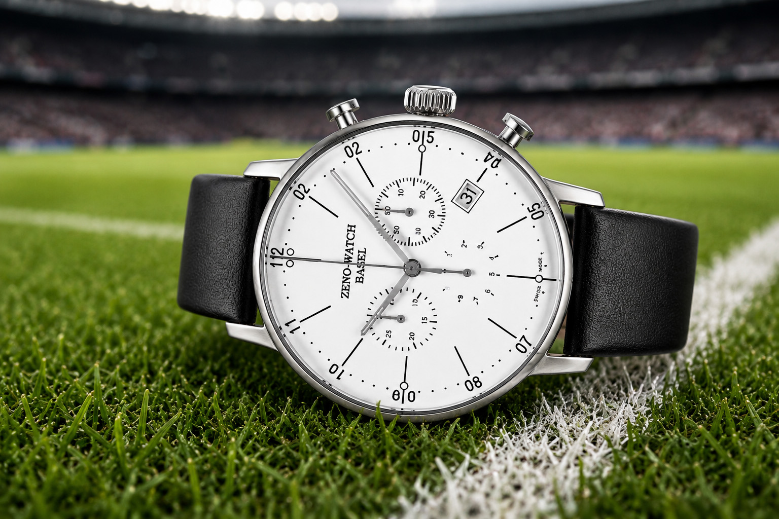 Close-up of a silver chronograph watch with a white face and black leather strap on a grassy sports field.